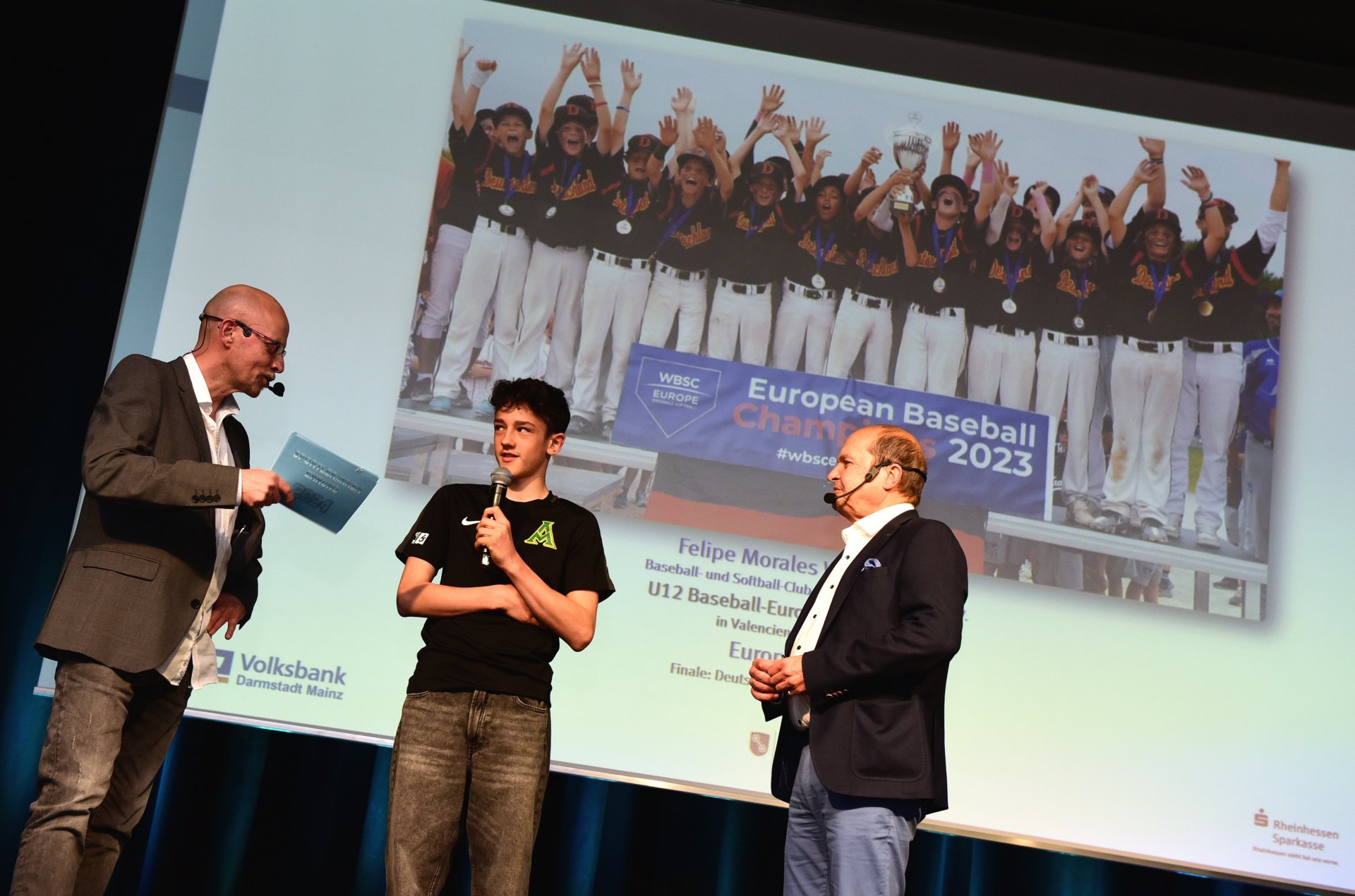 „Der Kleine mit dem harten Schlag“ lautete vor einem Jahr die Zeile in Anerkennung der Leistung von Felipa Morales Weggemann. Damals war der Baseballer der Mainz Athletics für den EM-Titel mit der deutschen U-12-Nationalmannschaft ausgezeichnet worden. Jetzt gab es einen Nachschlag: Auch 2023 reüssierte der junge Mainzer (hier zwischen Moderator Peter H. Eisenhuth und dem Paten Klaus Kuhn, Präsident des Sportbunds Rheinhessen) mit dem Team, diesmal mit 3:2 im Finale gegen die Niederlande, zu dem Felipe einen Run im ersten Inning beisteuerte.